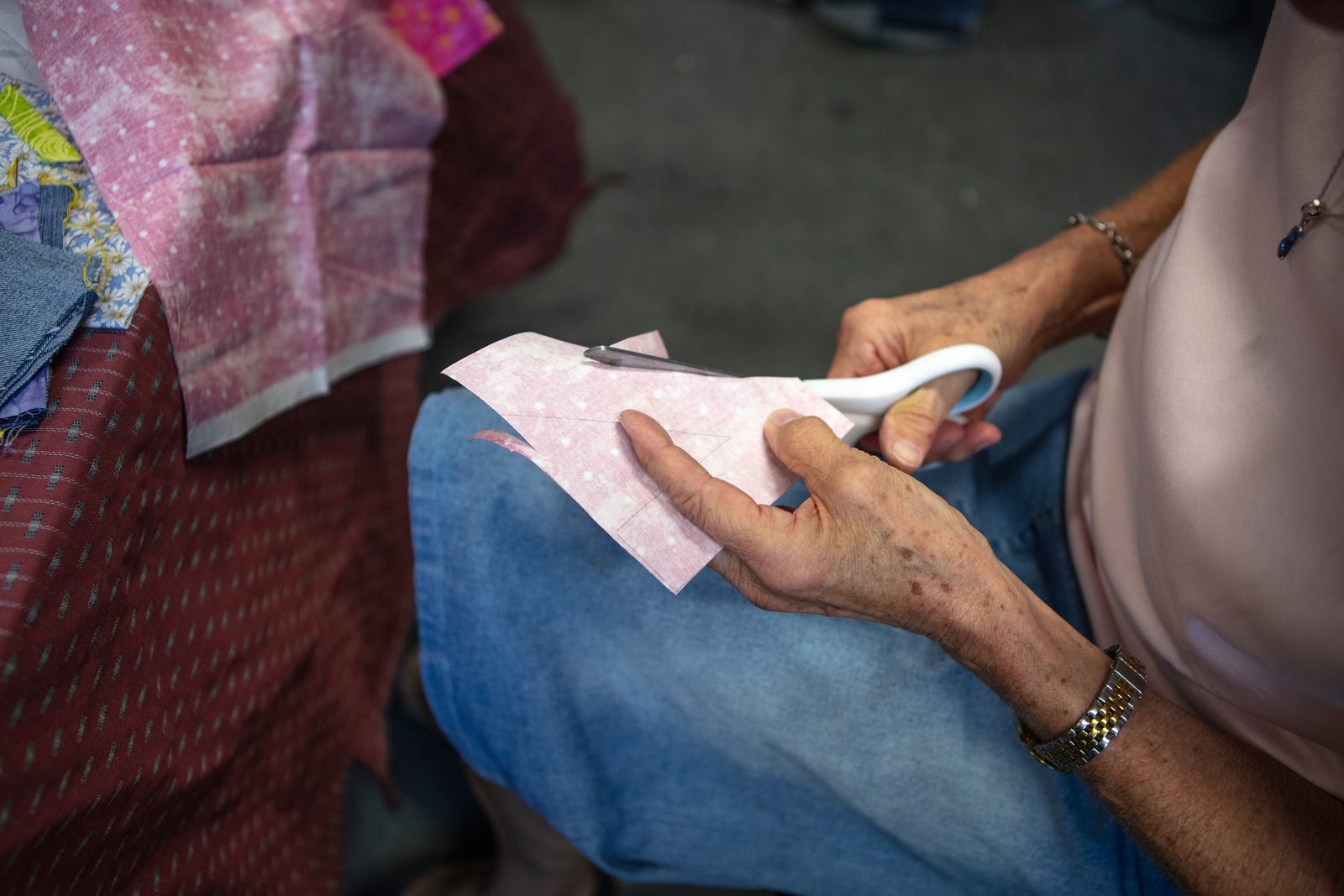 A participant cutting pink fabric on a denim square resting on their lap.