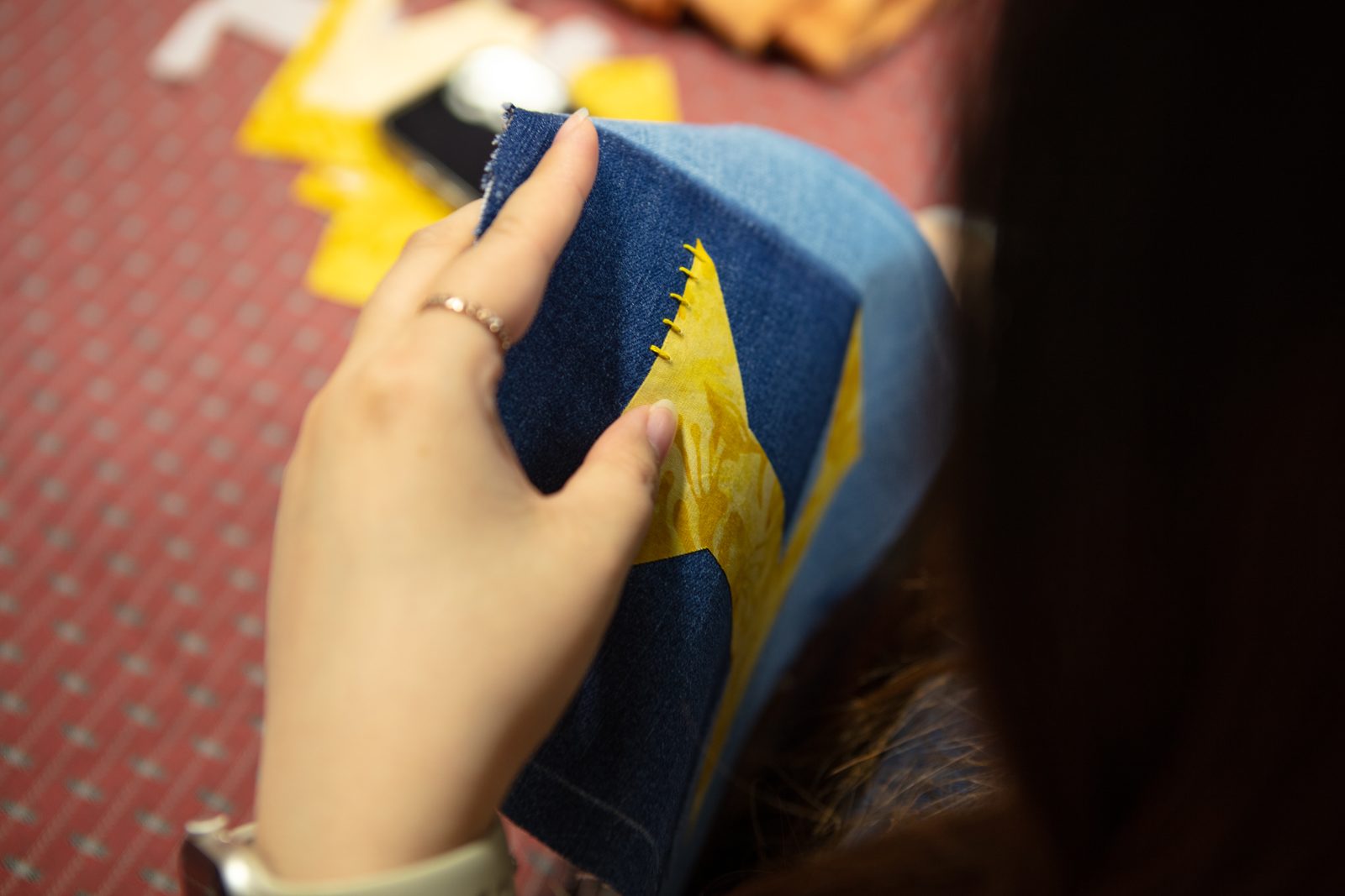A close-up of a hand stitching a yellow patch onto a denim square.