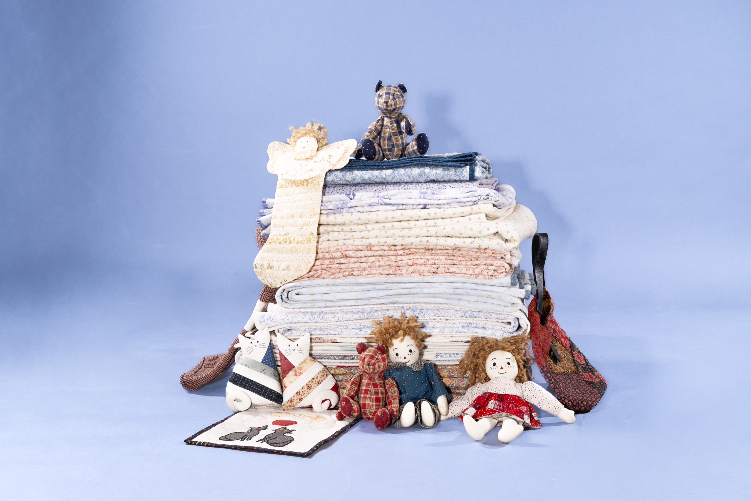 A tall stack of folded quilts topped with a handmade teddy bear and angel, surrounded by rag dolls, cloth cats and a woven bag, on a light blue backdrop.