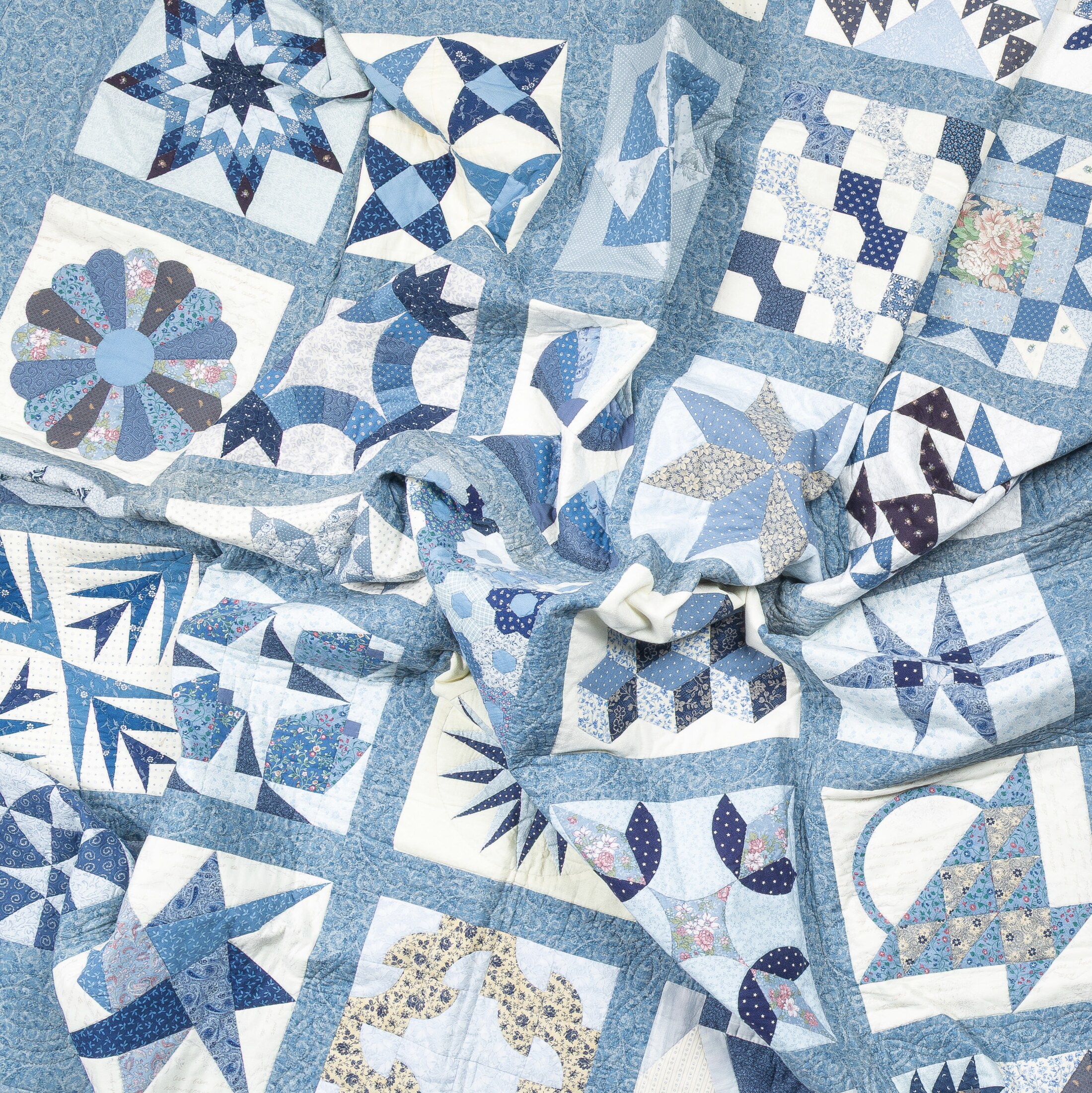 The full sampler quilt seen from above, bunched and crumpled at its center so the grid of blue star and pinwheel blocks folds in on itself.