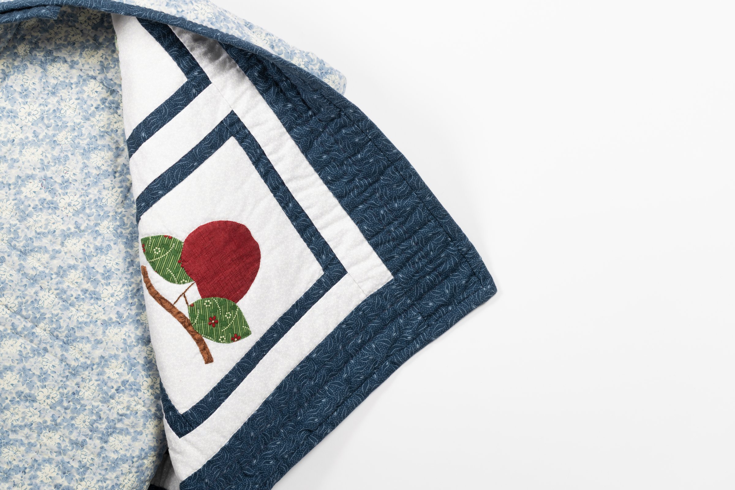 The folded corner of a quilt with an appliqued apple and leaves, its floral backing visible underneath.