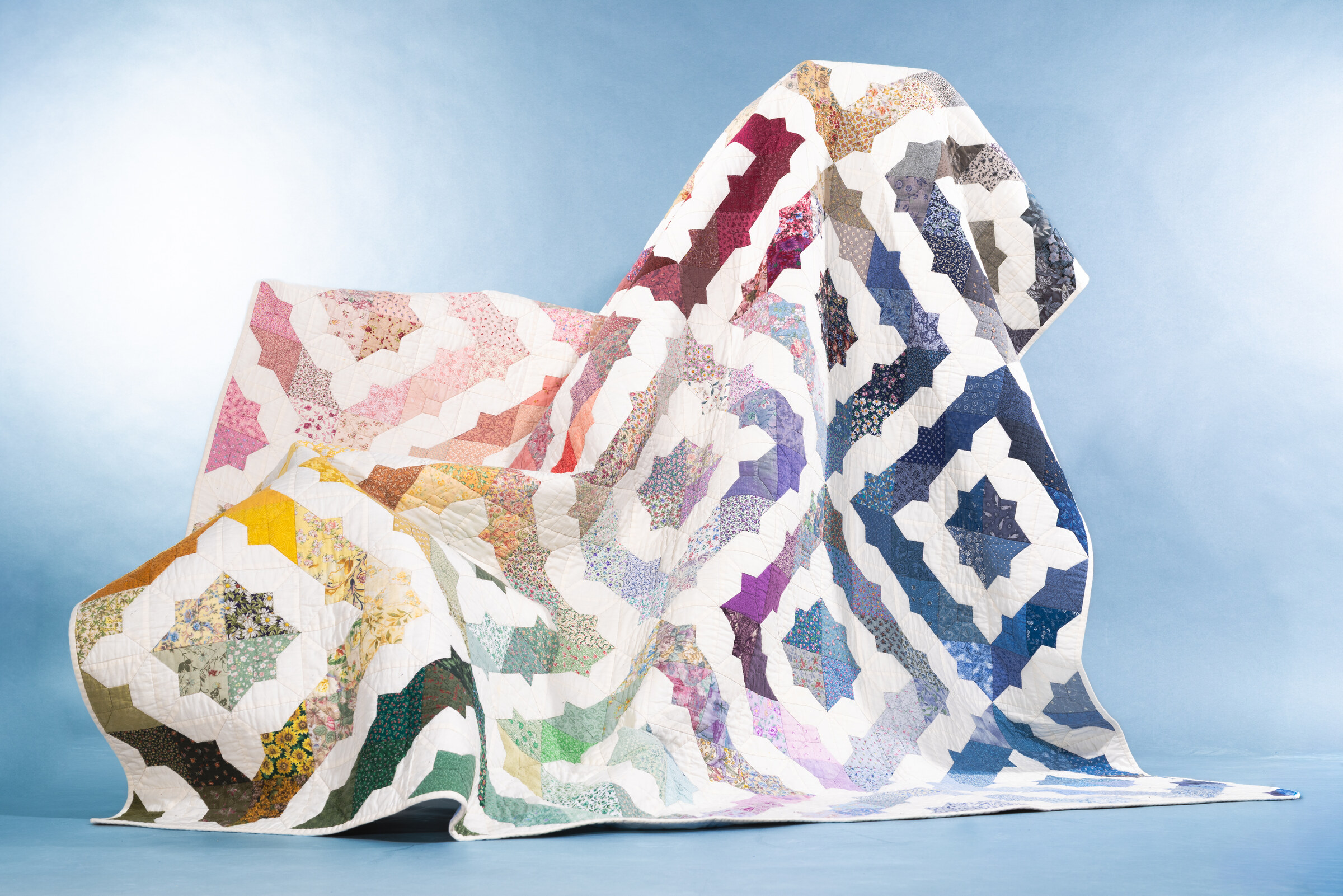 A large patchwork quilt in pink, yellow, green, purple and blue star blocks, draped loosely over a stand against a pale blue backdrop.