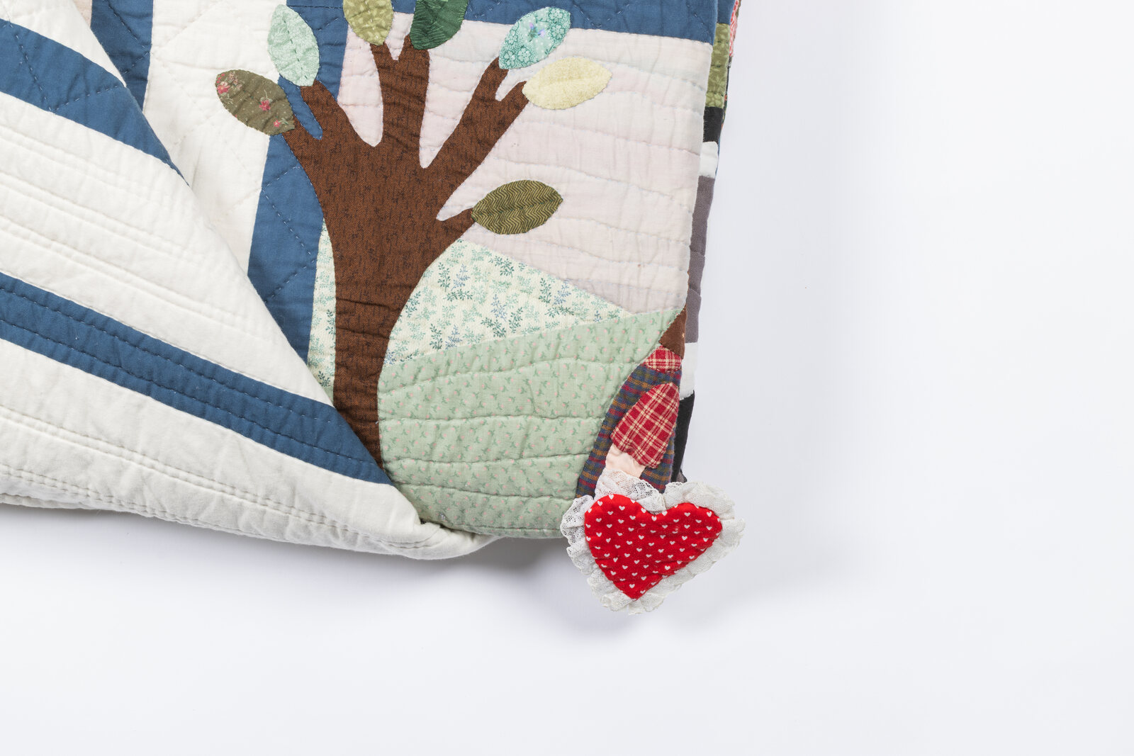 A folded quilt corner with an appliqued tree, a tiny appliqued figure and a red heart-shaped fabric patch trimmed with lace, photographed against a clean white studio backdrop.