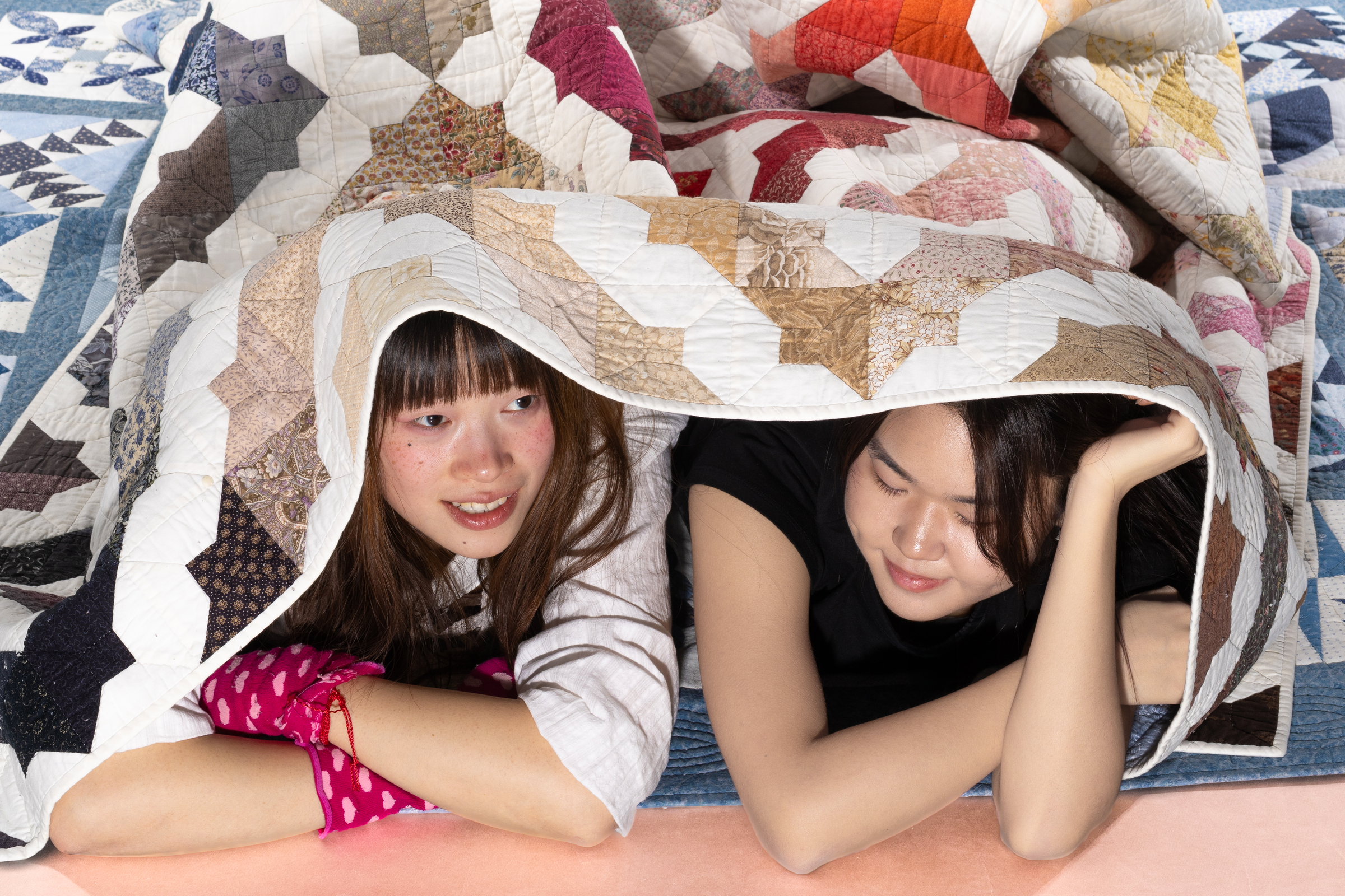 Two women lying together under a patchwork quilt lifted like a tent over their heads, surrounded by more folded quilts.