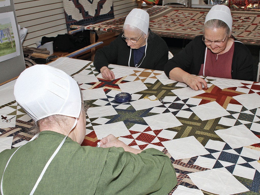 Three Amish quilters in bonnets hand-stitching a star-pattern quilt together at a table.