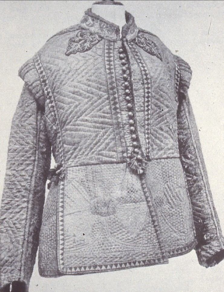 A black-and-white photograph of a quilted gambeson jacket with chevron-stitched sleeves and torso and a woven diamond-pattern skirt, displayed on a mannequin.