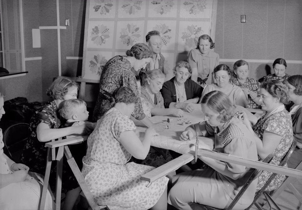 A black-and-white historical photograph of women and children gathered around a quilting frame.