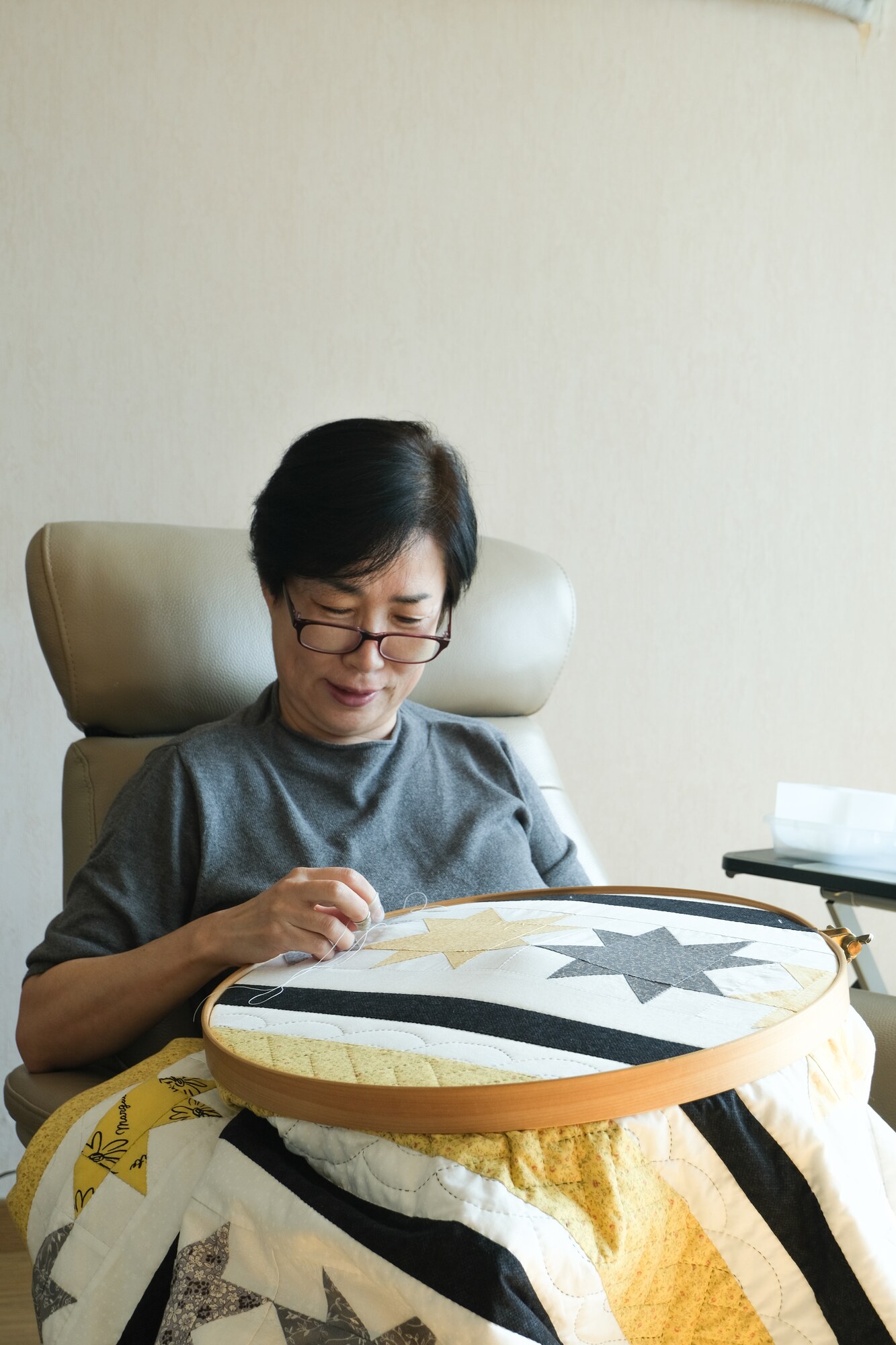 Kim Minseon hand-quilting a star-pattern quilt in a chair, wearing reading glasses.