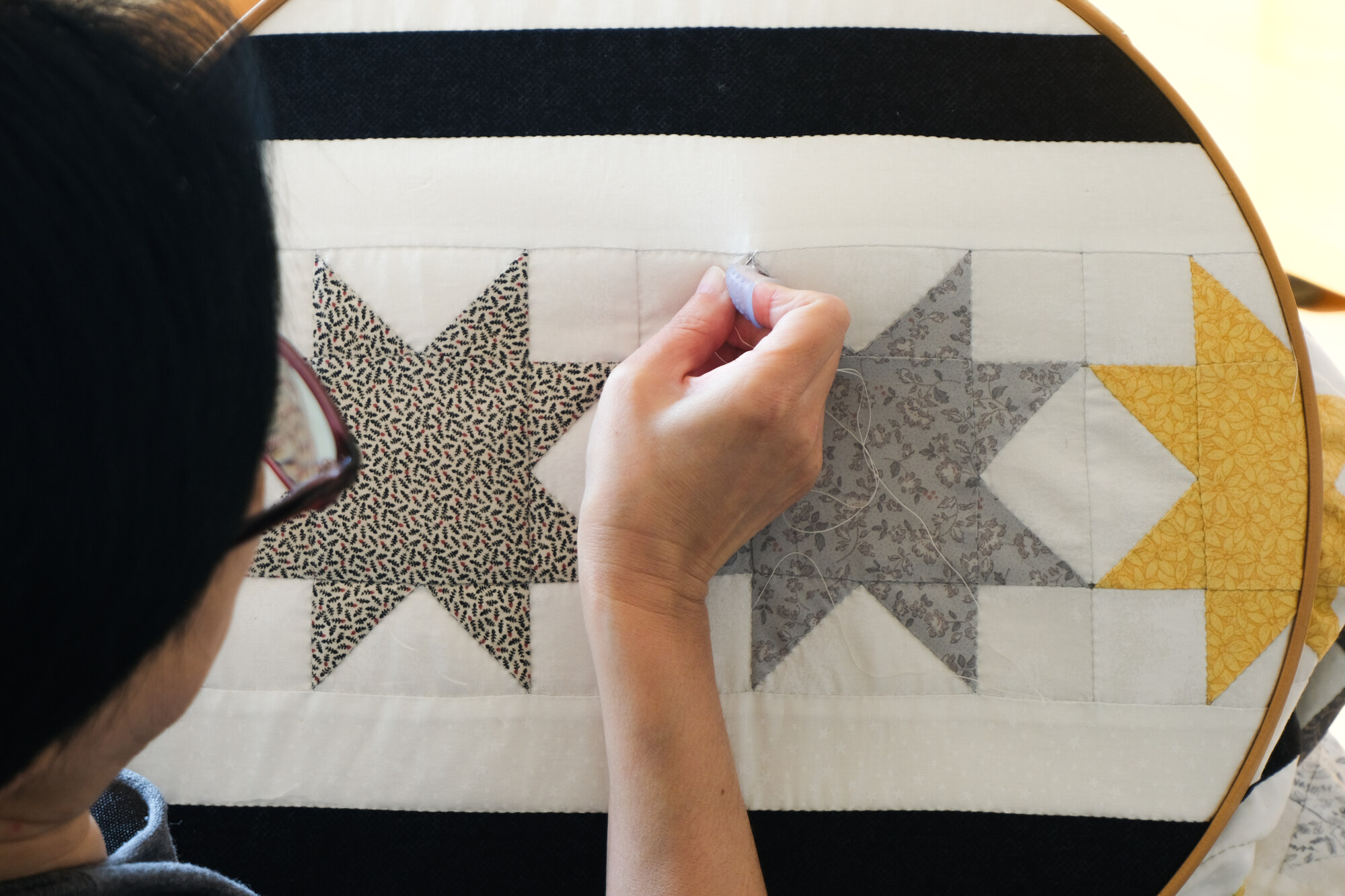 Close, over-the-shoulder view of Kim Minseon stitching a black, white and gold star-pattern quilt held in a hoop.