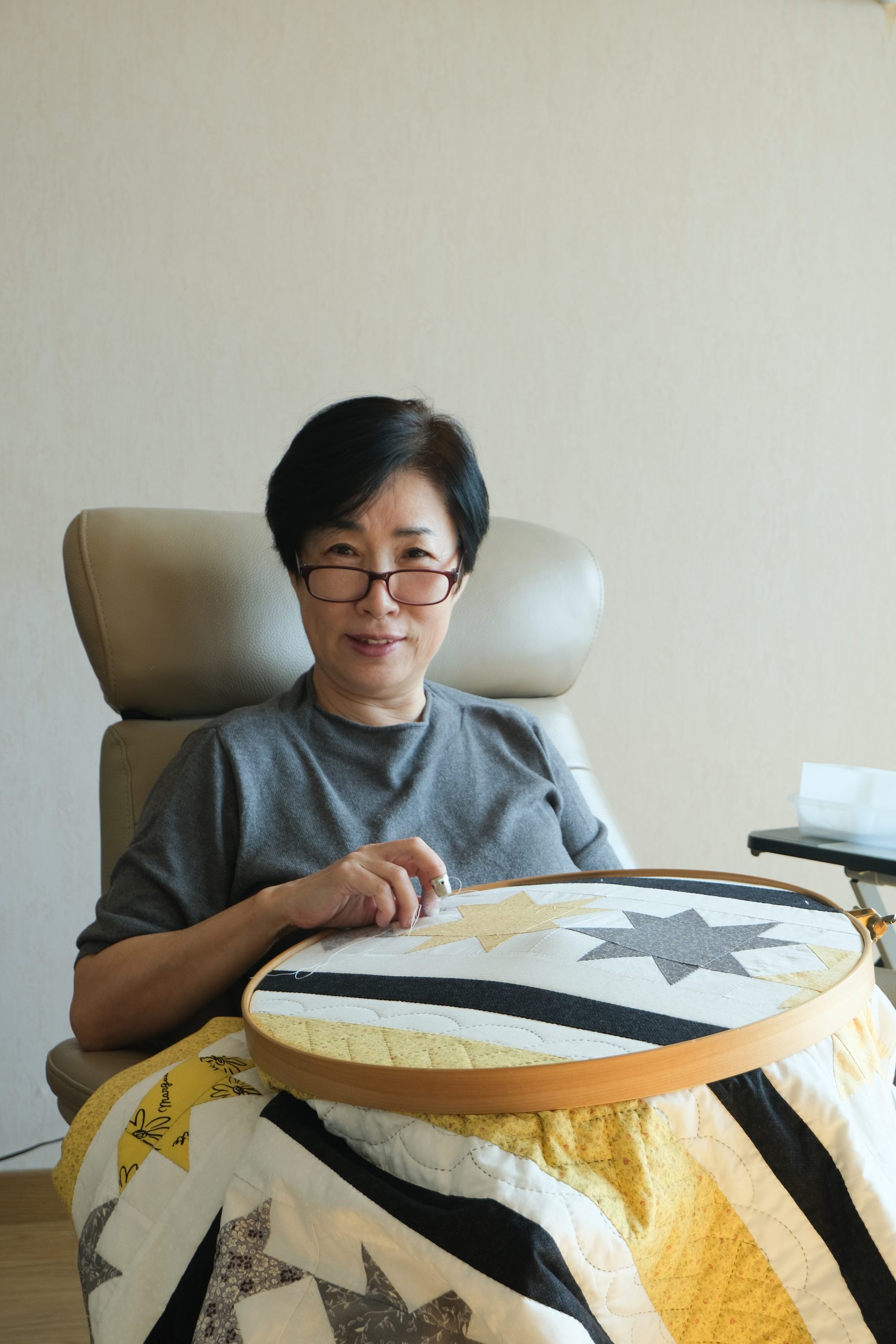 Kim Minseon sitting in a chair by the window, hand quilting a yellow, grey and white star-pattern quilt in a hoop.